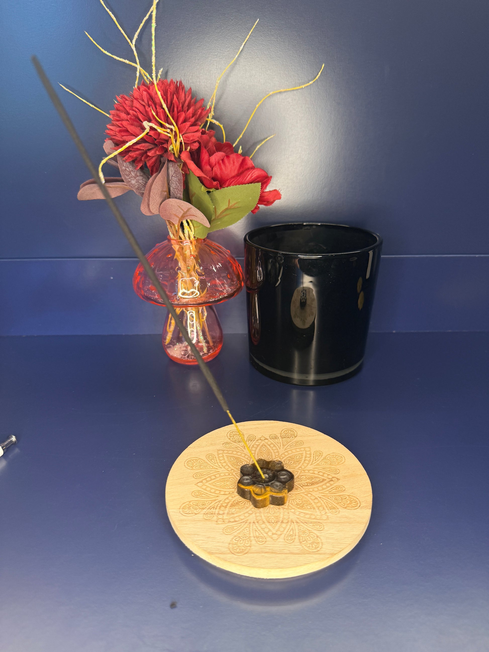 Tiger's Eye sunflower holding an incense stick while on a wooden coaster in front of a red flower and black jar on a blue background