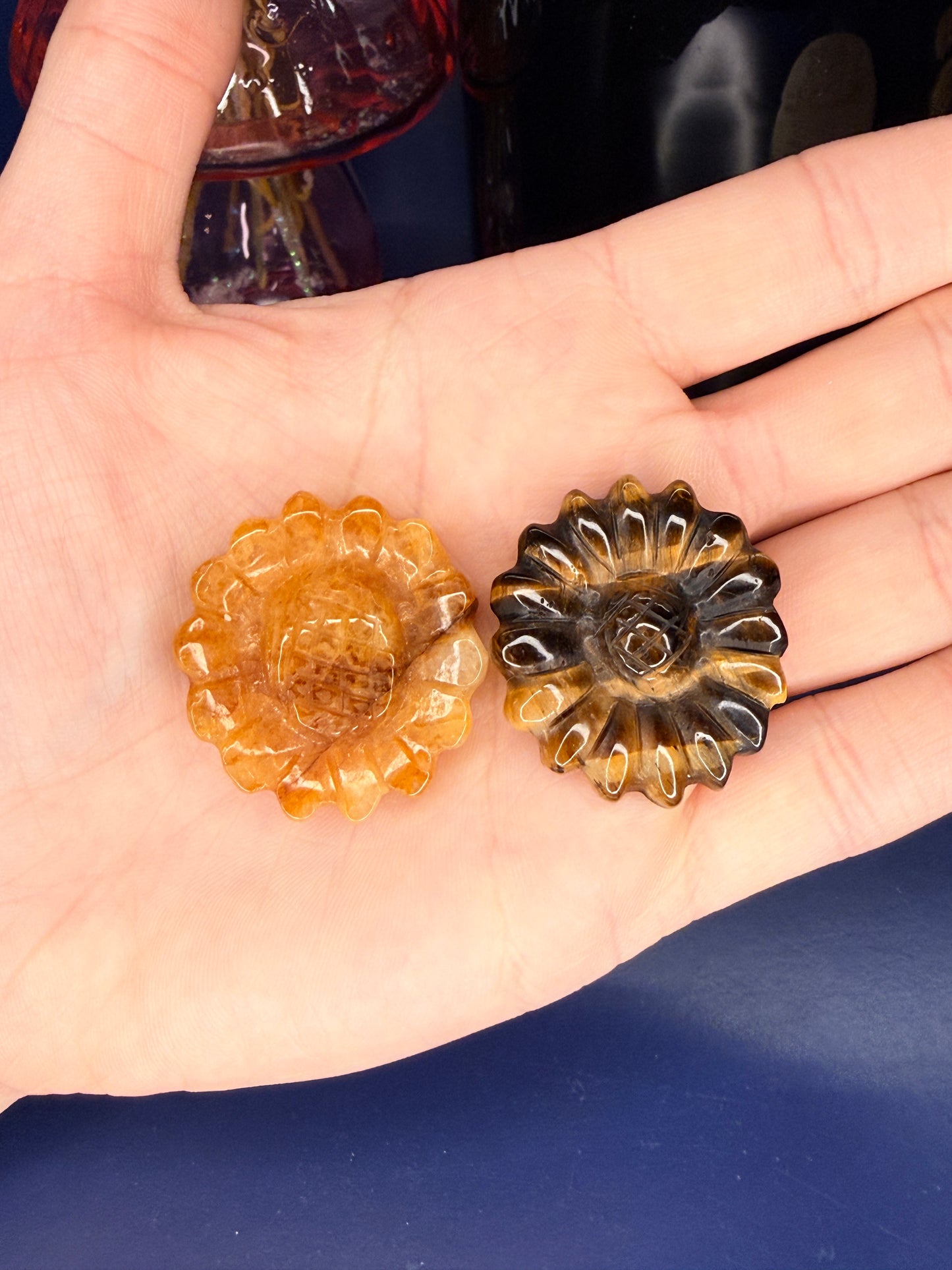 Hand holding orange and golden crystal flowers in front of a pink vase and black jar on a blue background