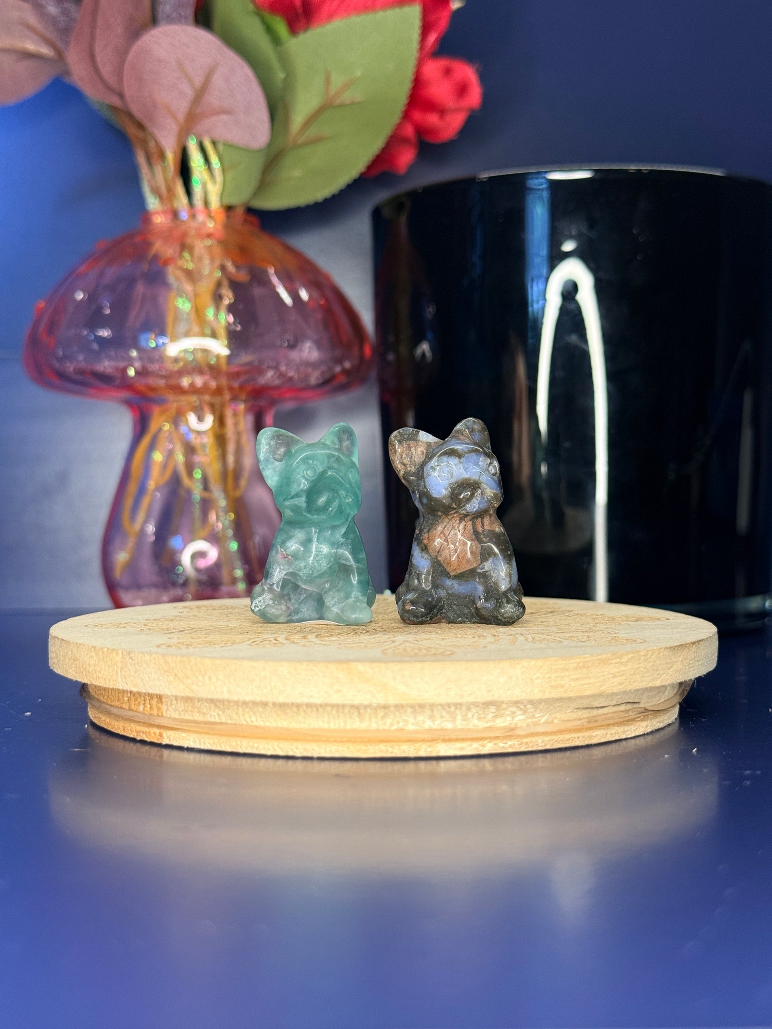 Green and multicolored dog carvings in front of a pink vase and black jar on a wooden coaster against a blue background
