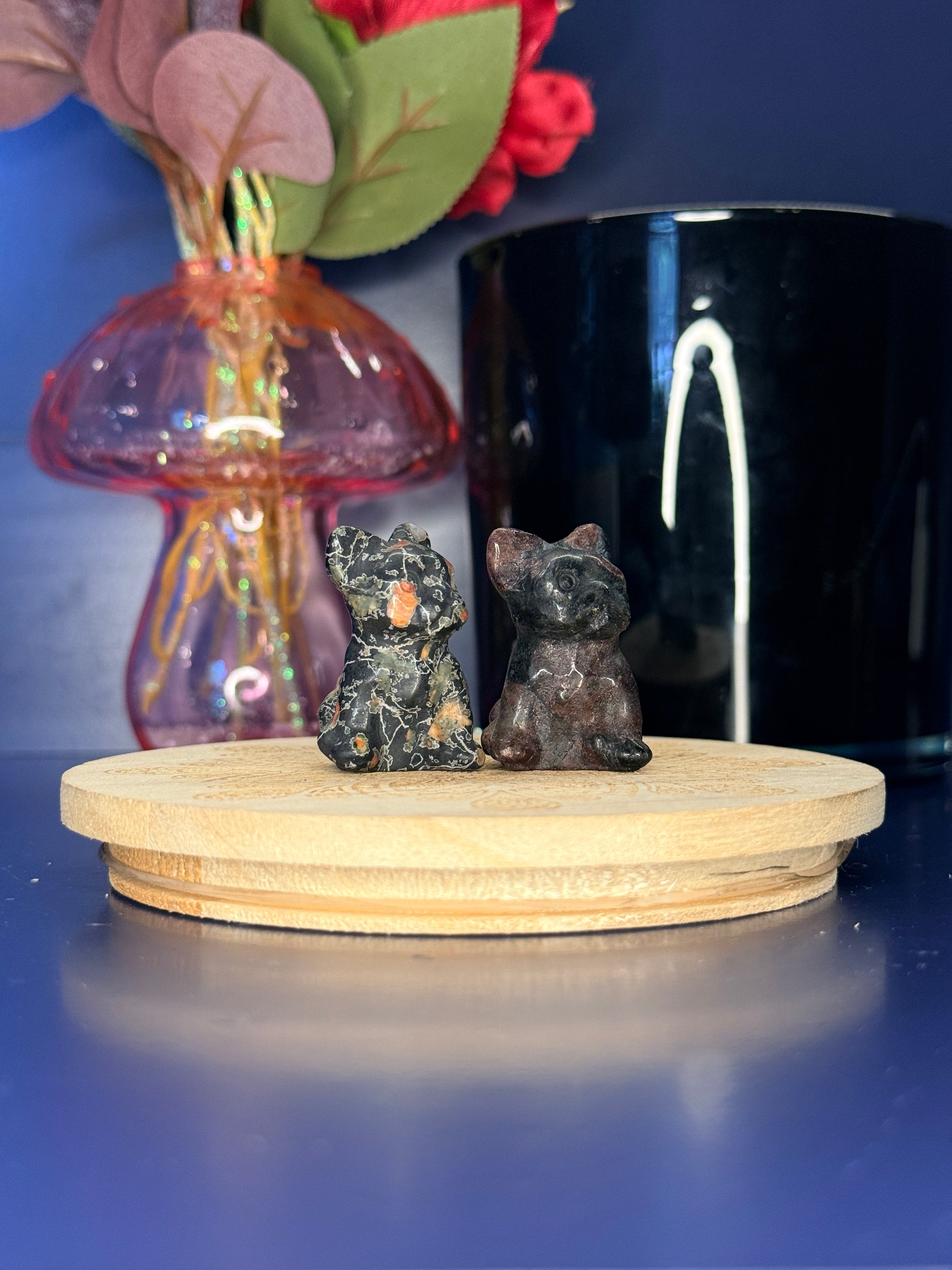 Two multicolored crystal dog carvings in front of a pink vase and black jar on a wooden coaster against a blue background
