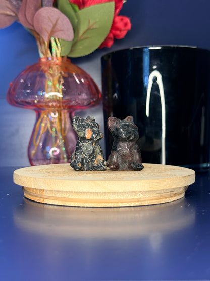 Two multicolored crystal dog carvings in front of a pink vase and black jar on a wooden coaster against a blue background