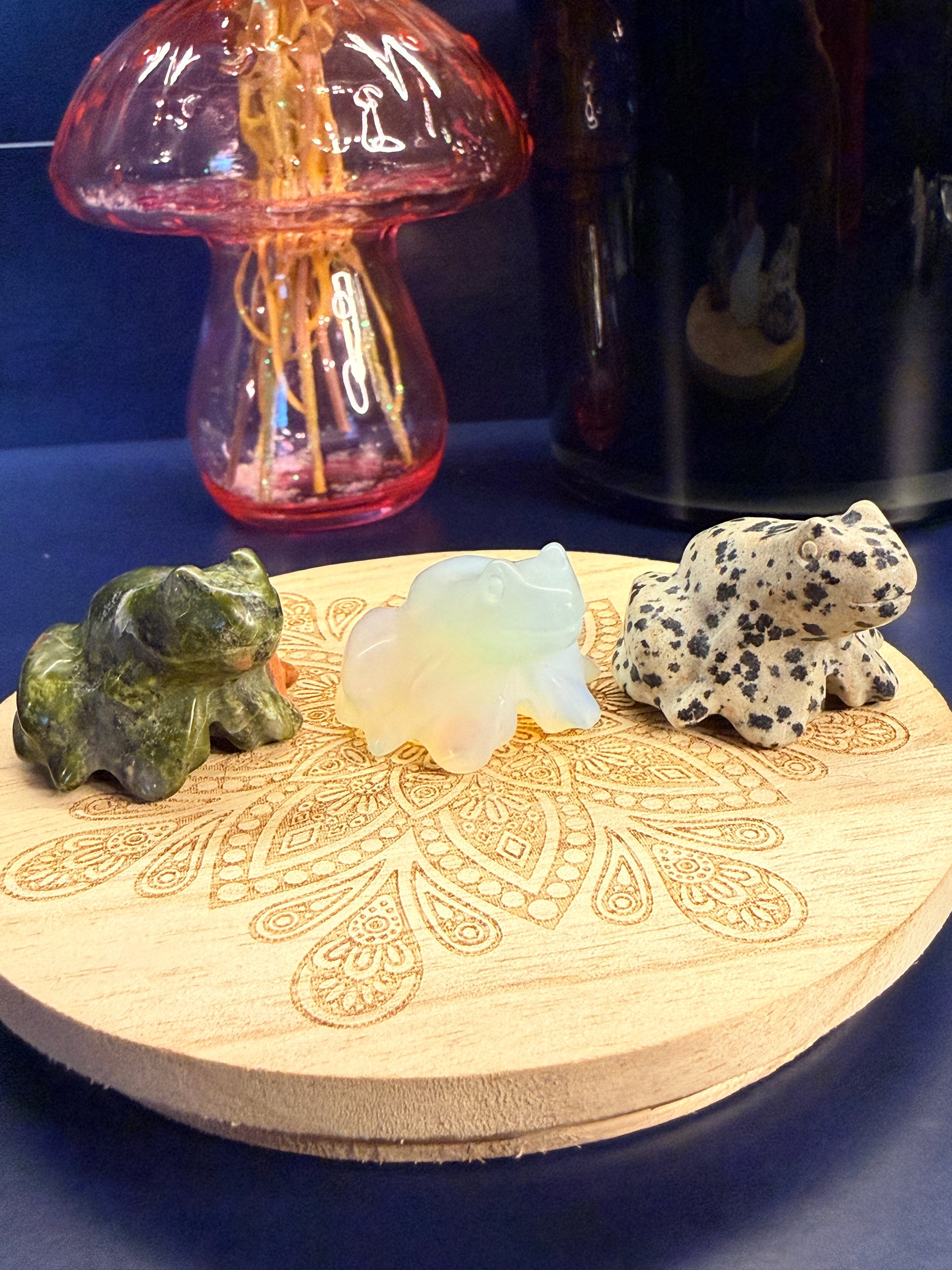 Three crystal frogs sitting on a decorative wooden coaster in front of a glass mushroom and black jar