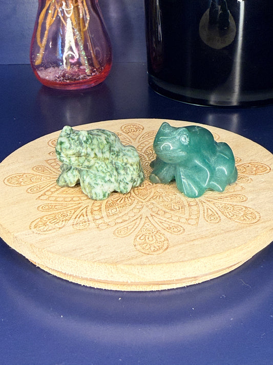 Two green crystal frog carvings in front of a pink vase and black jar on a wooden coaster against a blue background