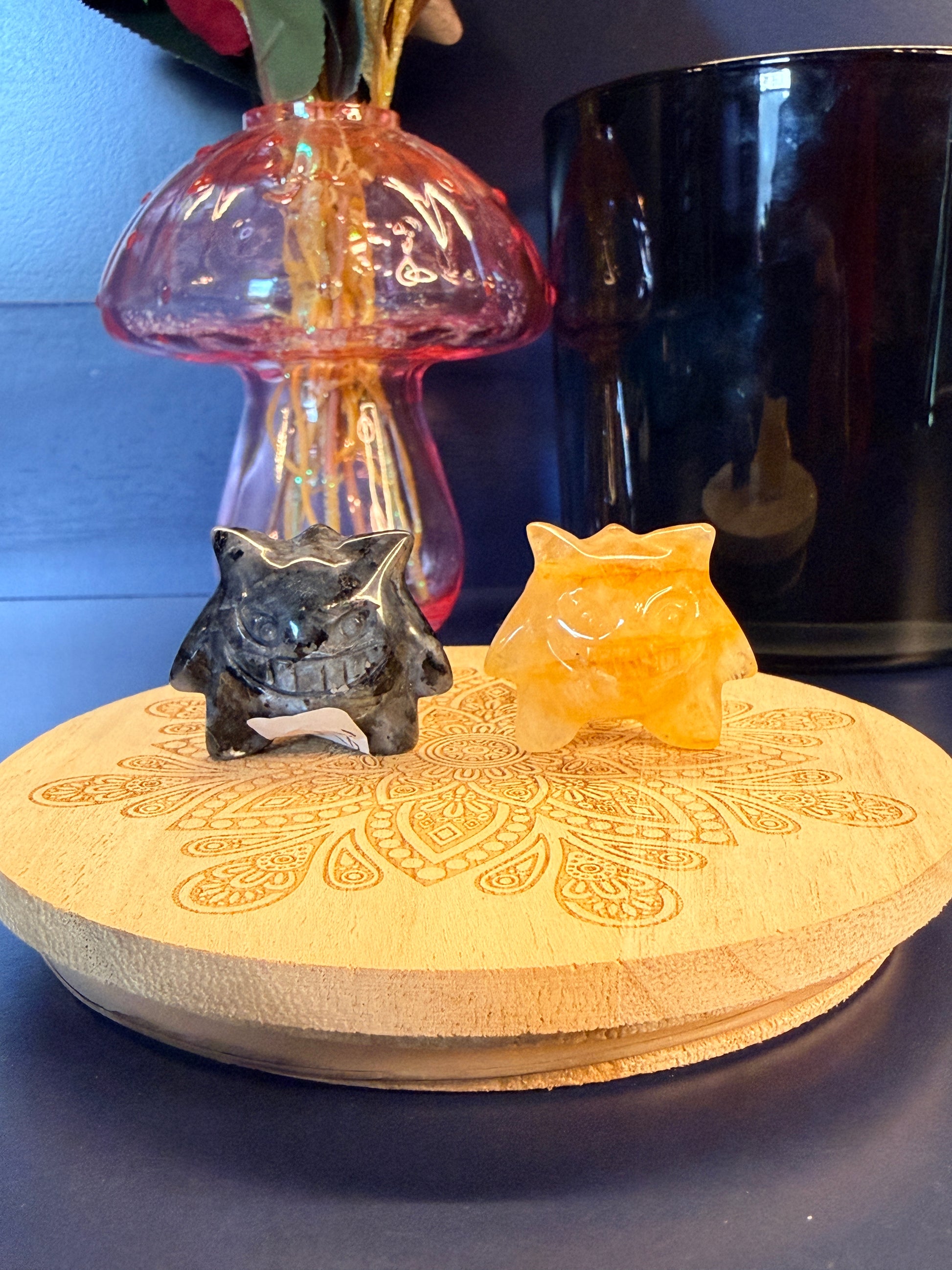 Two gengar-inspired crystals, larvikite and golden healer quartz, sitting on a wooden coaster in front of a glass mushroom and black jar