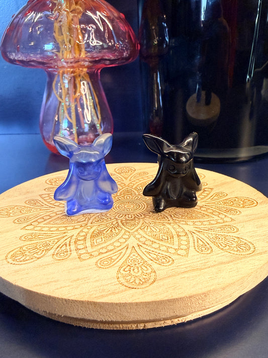 Two different crystal glaceons sitting on a decorative wooden coaster in front of a glass mushroom and black jar