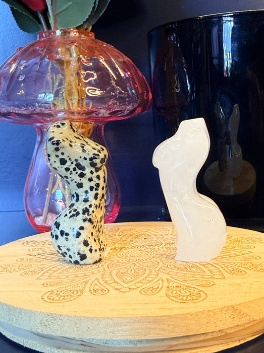 Two goddess bodies, one spotted and one white, sitting on a decorative wooden coaster in front of a mushroom and black jar