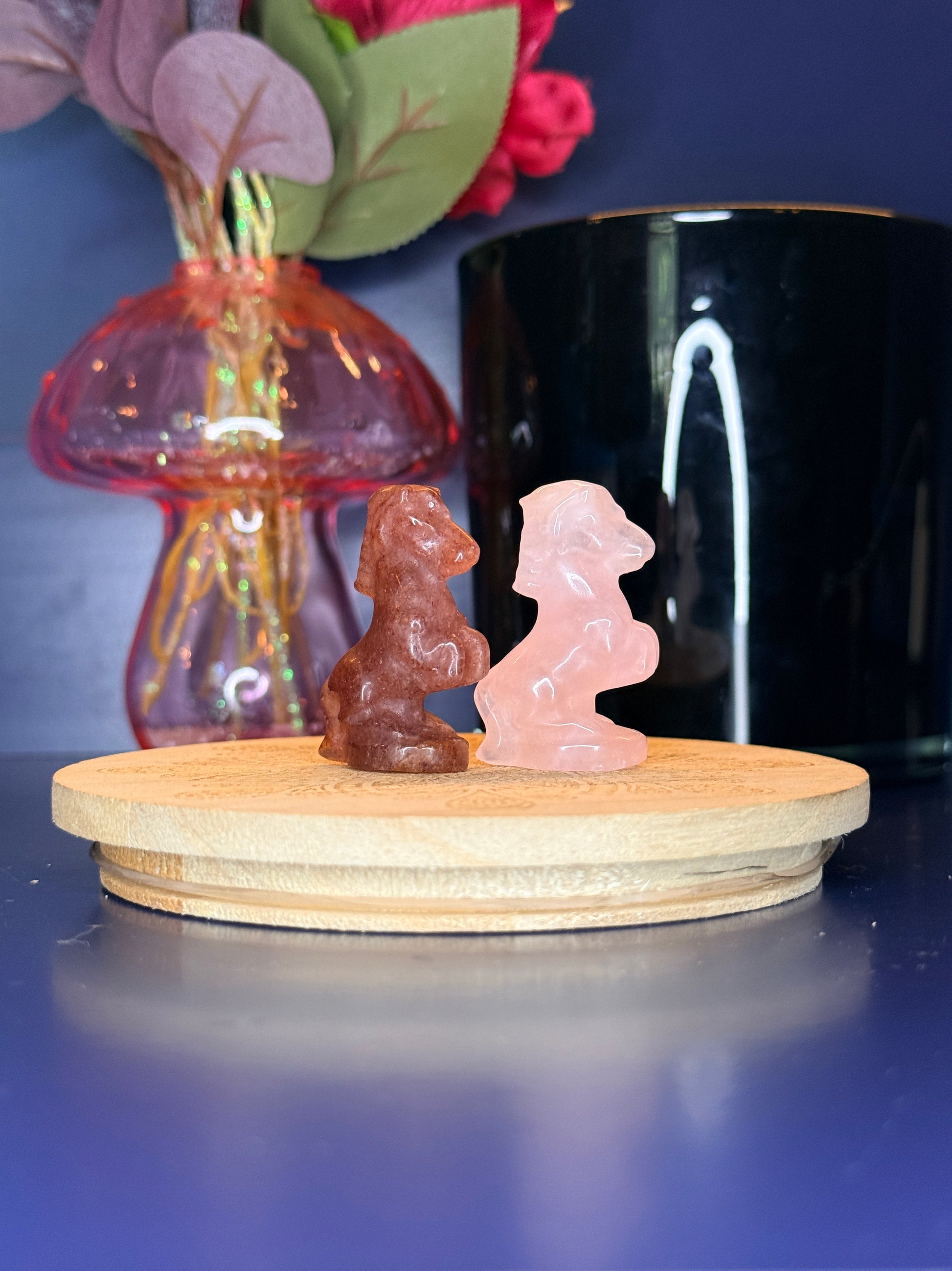 Red and pink crystal horse carvings in front of a pink vase and black jar on a wooden coaster against a blue background