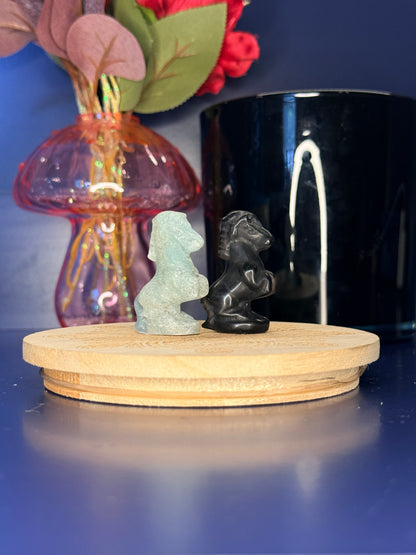 Blue and black crystal horse carvings in front of a pink vase and black jar on a wooden coaster against a blue background