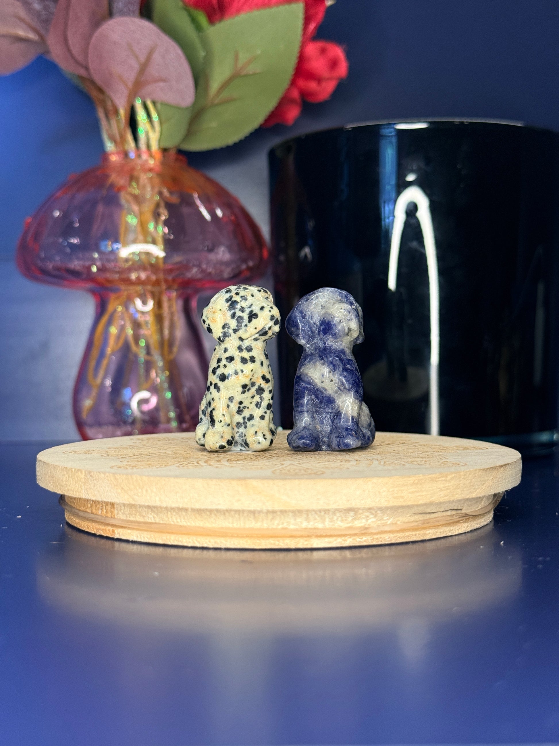 Spotted and blue crystal dog carvings in front of a pink vase and black jar on a wooden coaster against a blue background