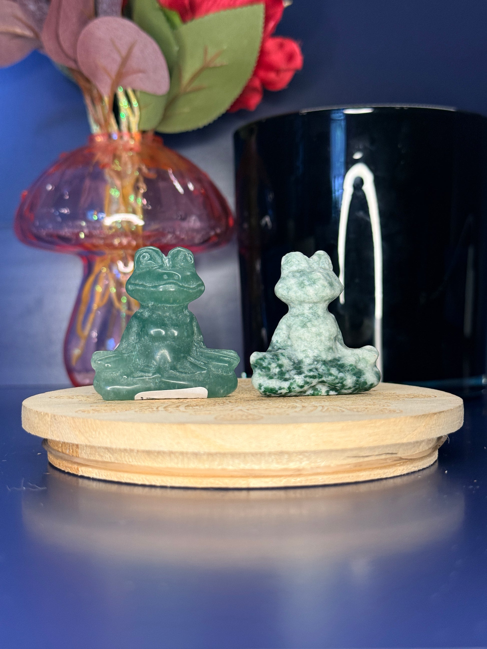 Green crystal meditating frog carvings in front of a pink vase and black jar on a wooden coaster against a blue background