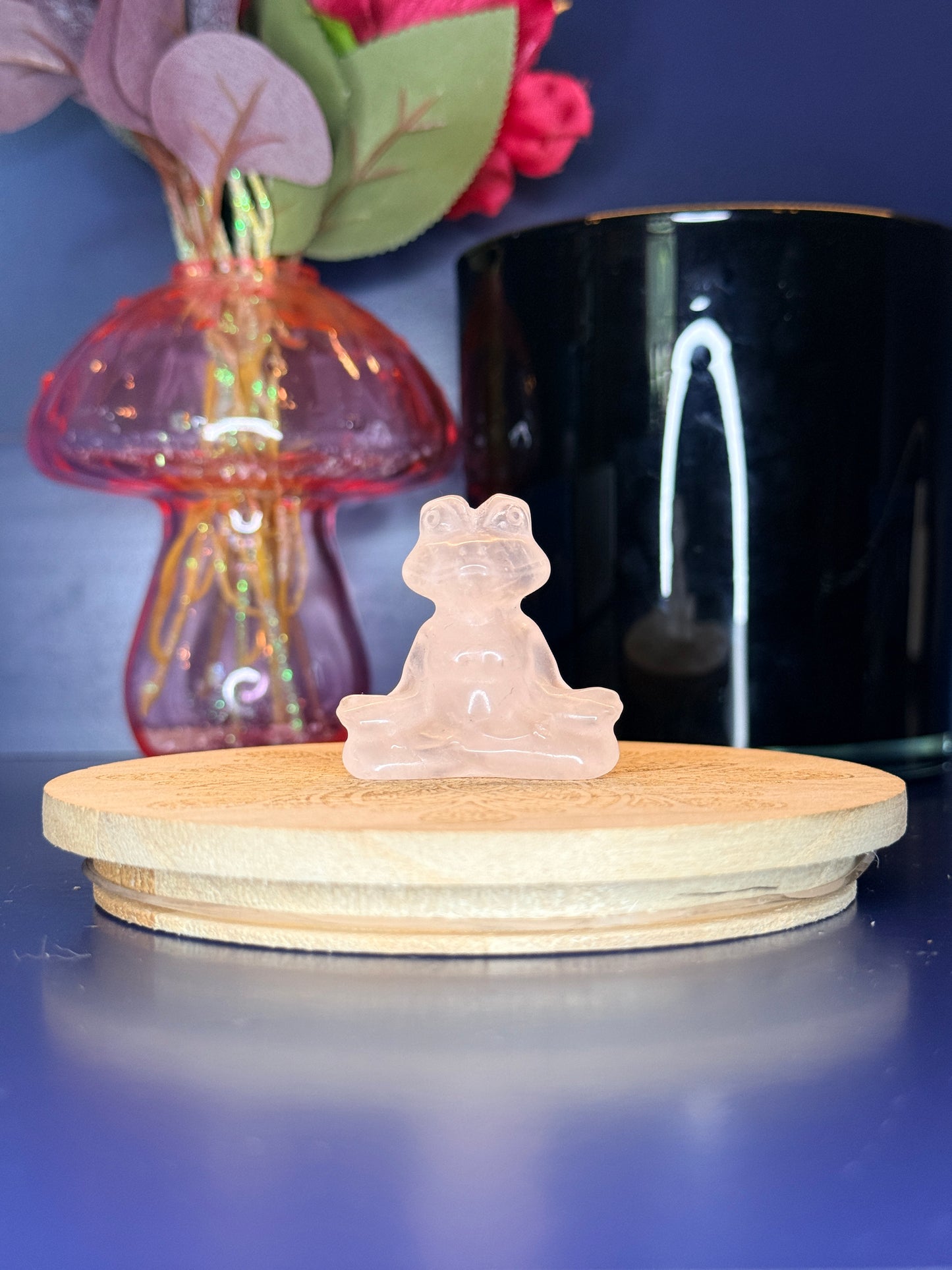 Pink rose quartz meditating frog carving in front of a pink vase and black jar on a wooden coaster against a blue background