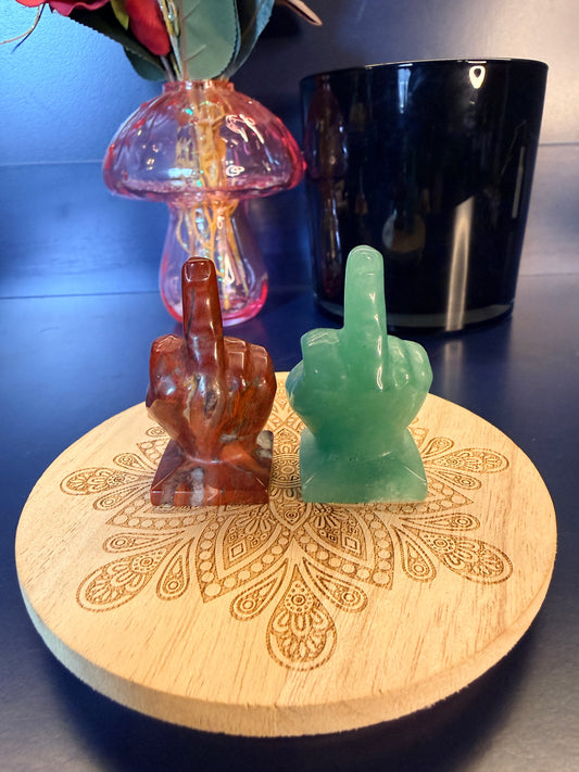 Two crystal middle fingers sitting on a decorative wooden coaster in front of a glass mushroom and black jar