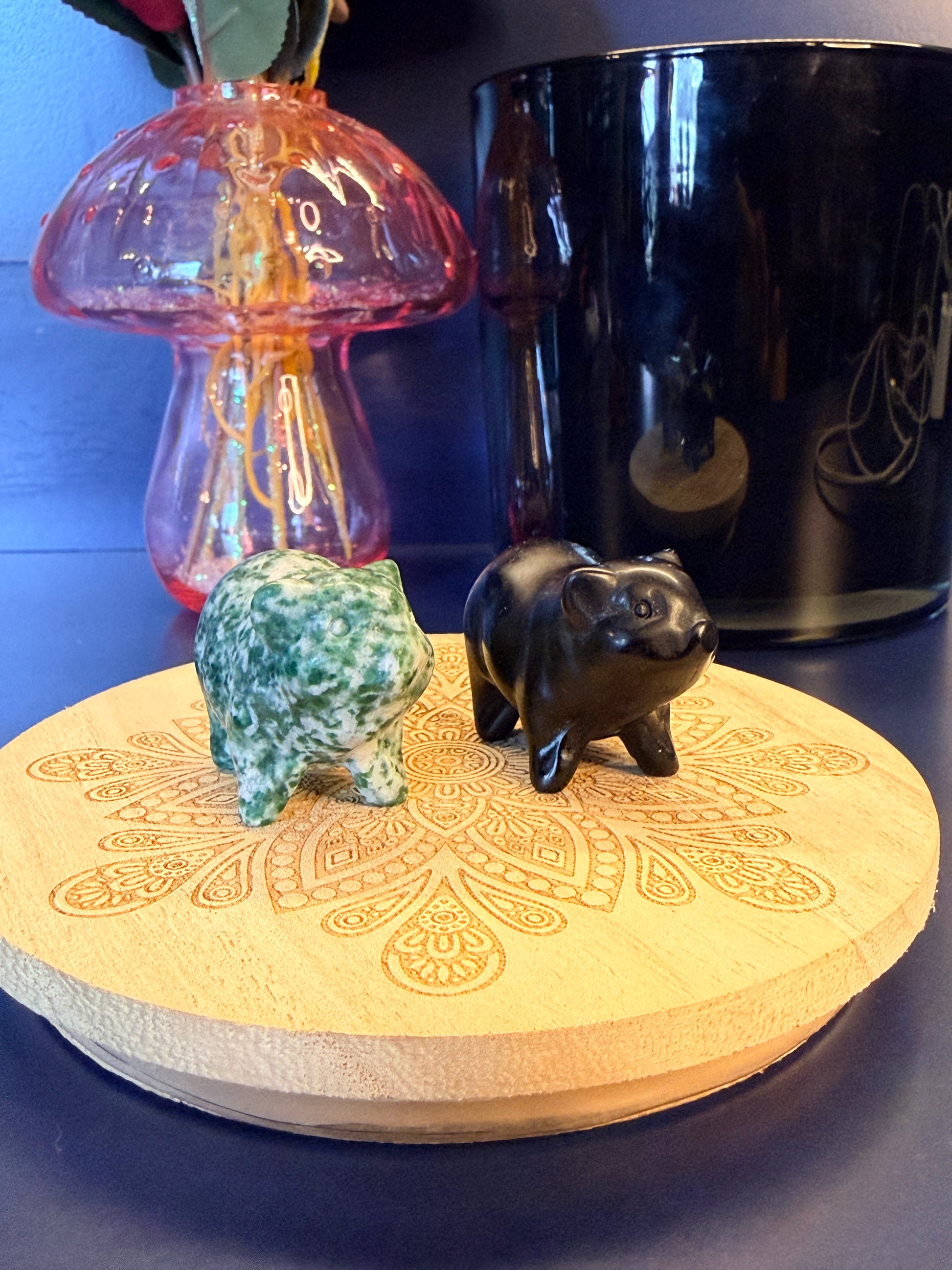 Two crystal pigs sitting on a decorative wooden coaster in front of a glass mushroom and black jar