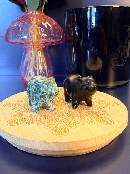 Two crystal pigs sitting on a decorative wooden coaster in front of a glass mushroom and black jar
