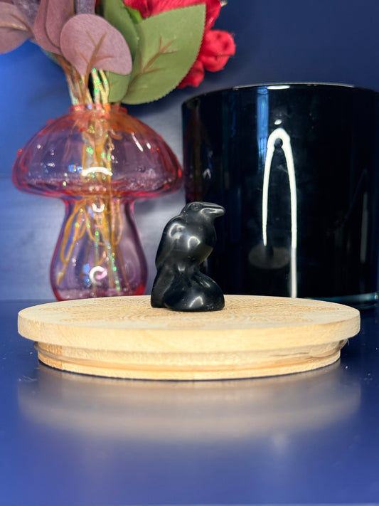 Black obsidian raven carving in front of a pink vase and black jar on a wooden coaster against a blue background