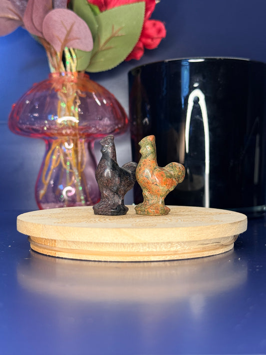 Multicolored crystal rooster carvings in front of a pink vase and black jar on a wooden coaster against a blue background