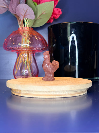 Strawberry quartz rooster carving in front of a pink vase and black jar on a wooden coaster against a blue background