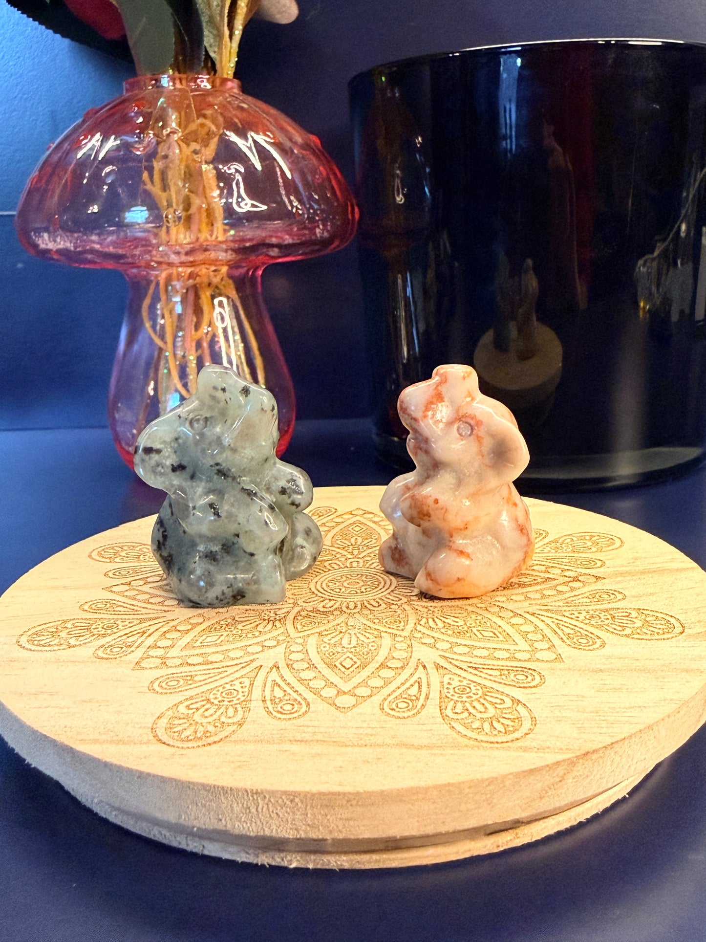 Two crystal sitting elephants sitting on a decorative wooden coaster in front of a glass mushroom and black jar