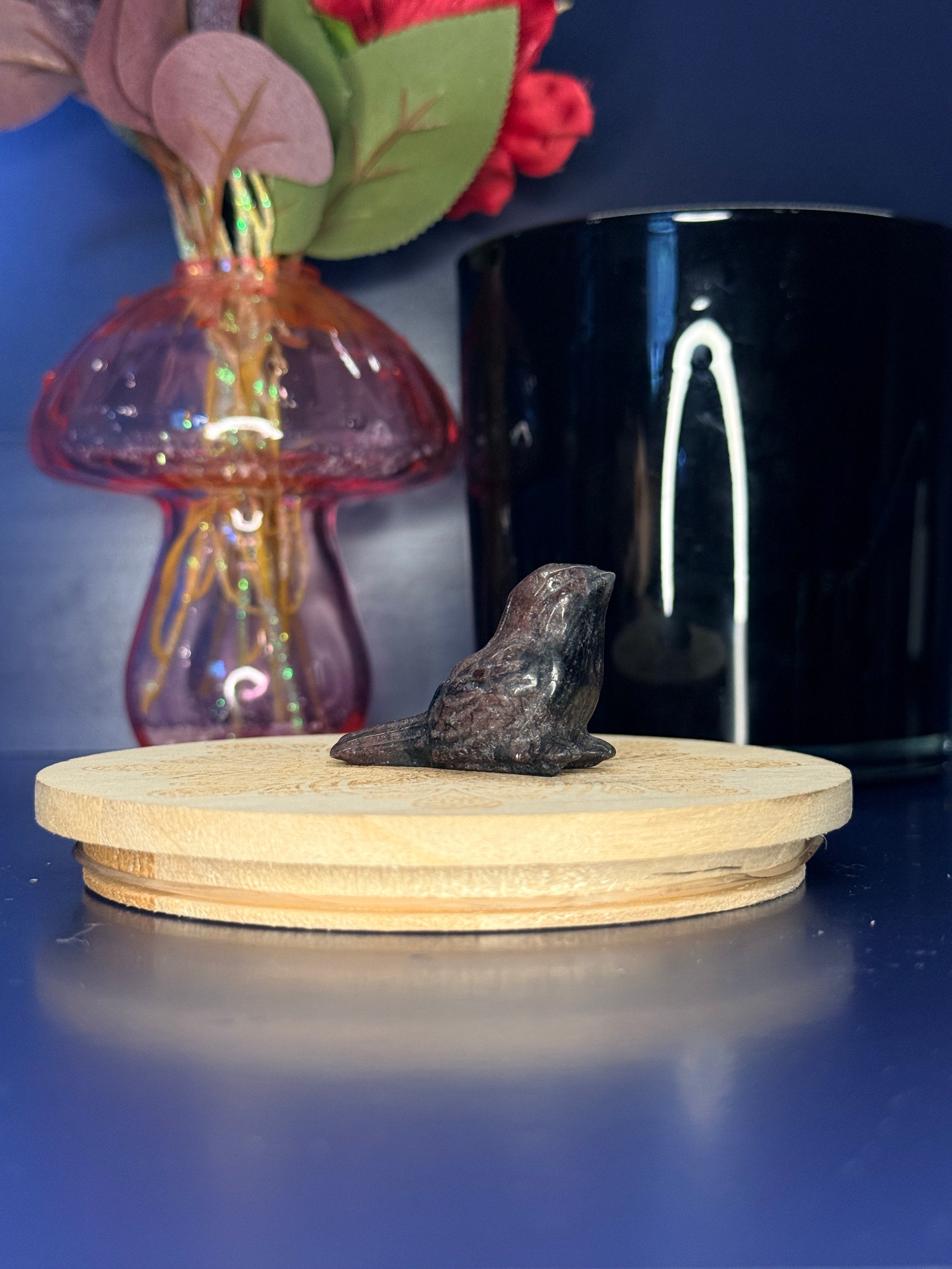 Red and black garnet sparrow carving in front of a pink vase and black jar on a wooden coaster against a blue background