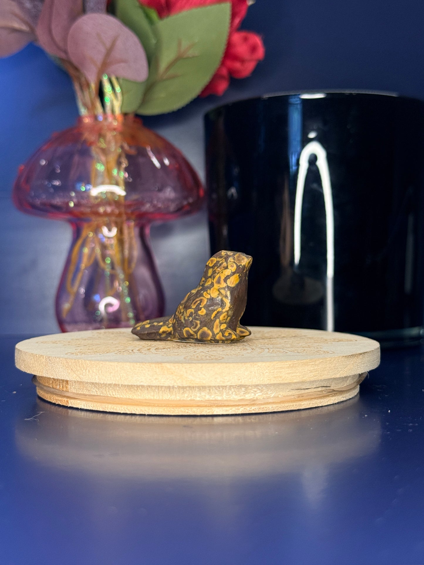 Yellow and brown fruit jasper sparrow carving in front of a pink vase and black jar on a wooden coaster against a blue background
