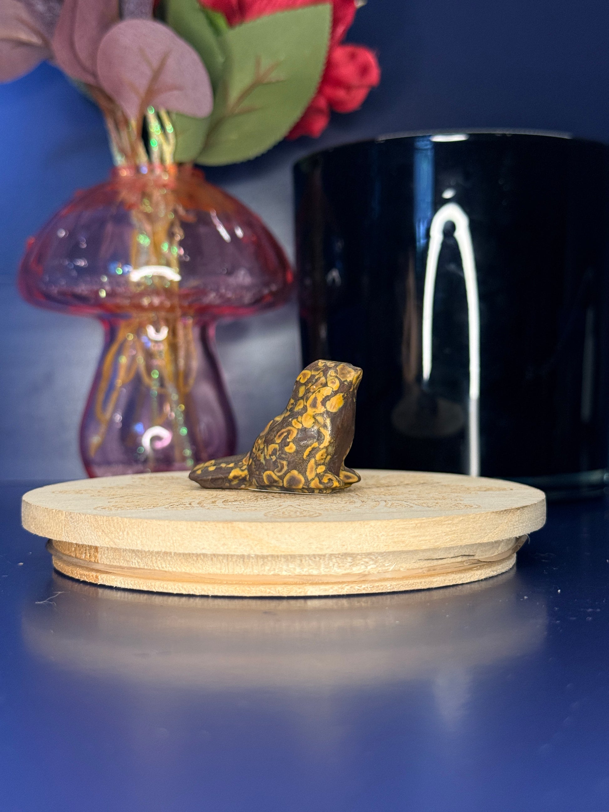 Yellow and brown fruit jasper sparrow carving in front of a pink vase and black jar on a wooden coaster against a blue background