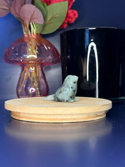 green and black kiwi jasper sparrow carving in front of a pink vase and black jar on a wooden coaster against a blue background