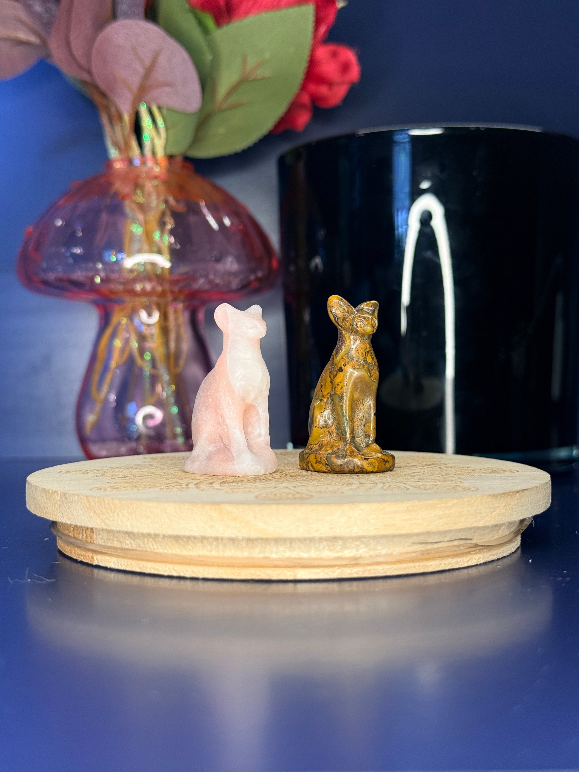 Pink and yellow sphinx cat carvings in front of a pink vase and black jar on a wooden coaster against a blue background