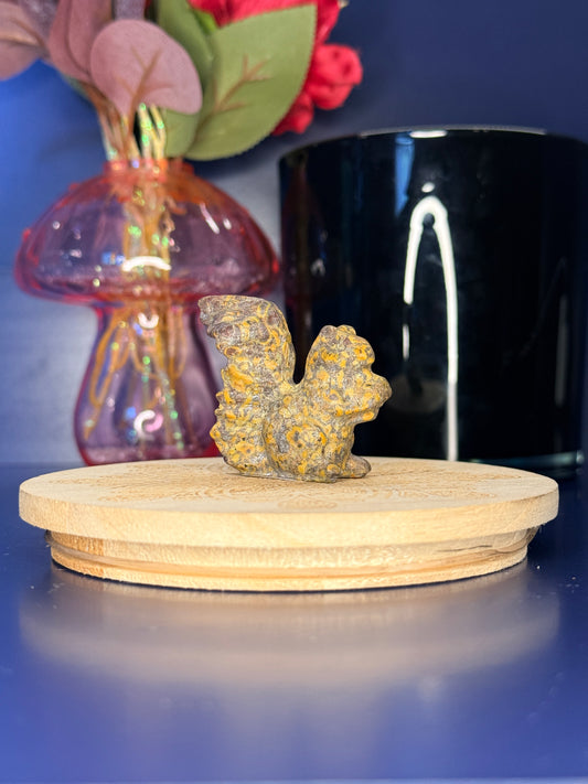 Multicolored fruit jasper squirrel carving in front of a pink vase and black jar on a wooden coaster against a blue background