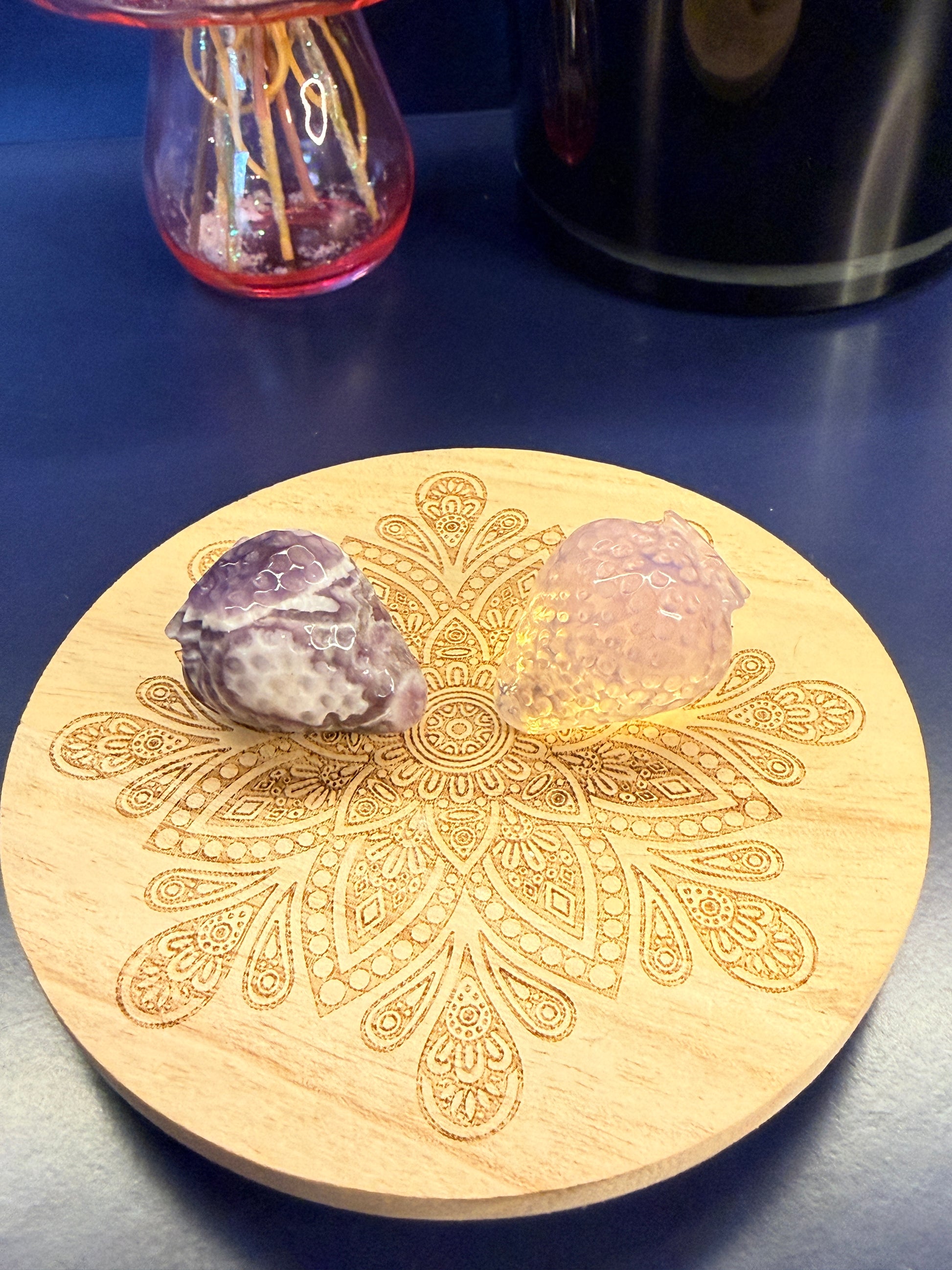 Two strawberry crystals sitting on a decorative wooden coaster in front of a glass mushroom and black jar