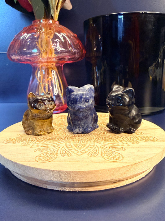 Three crystal cats sitting on a decorative wooden coaster in front of a glass mushroom and black jar