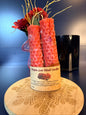 Two red candles in a decorative holder with a decorative wooden coaster and floral arrangement.