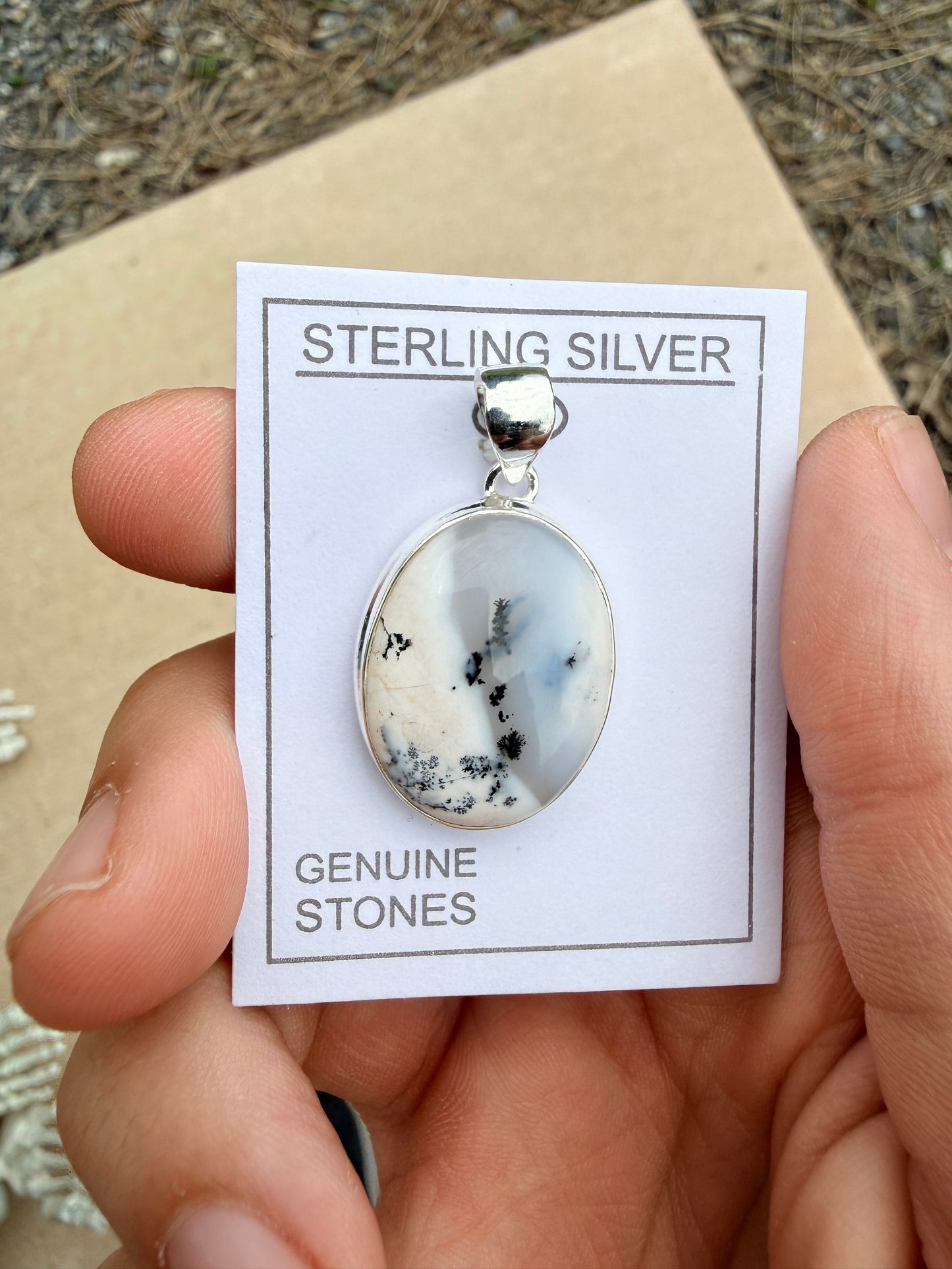 Hand holding a white and grey dendrite agate pendant on a white card against a blurred nature background
