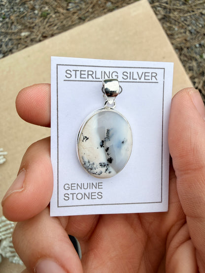 Hand holding a white and grey dendrite agate pendant on a white card against a blurred nature background