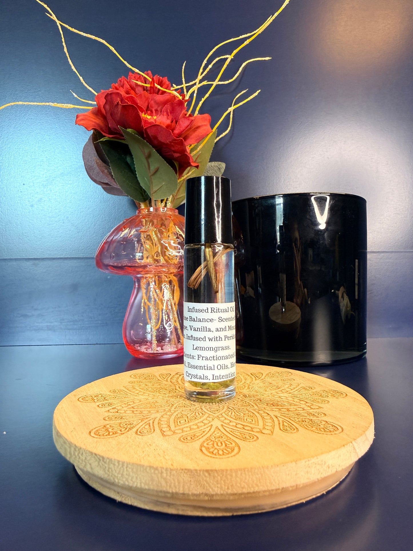 Body Oil Vial sitting on a decorative wooden coaster in front of a red flower and black jar