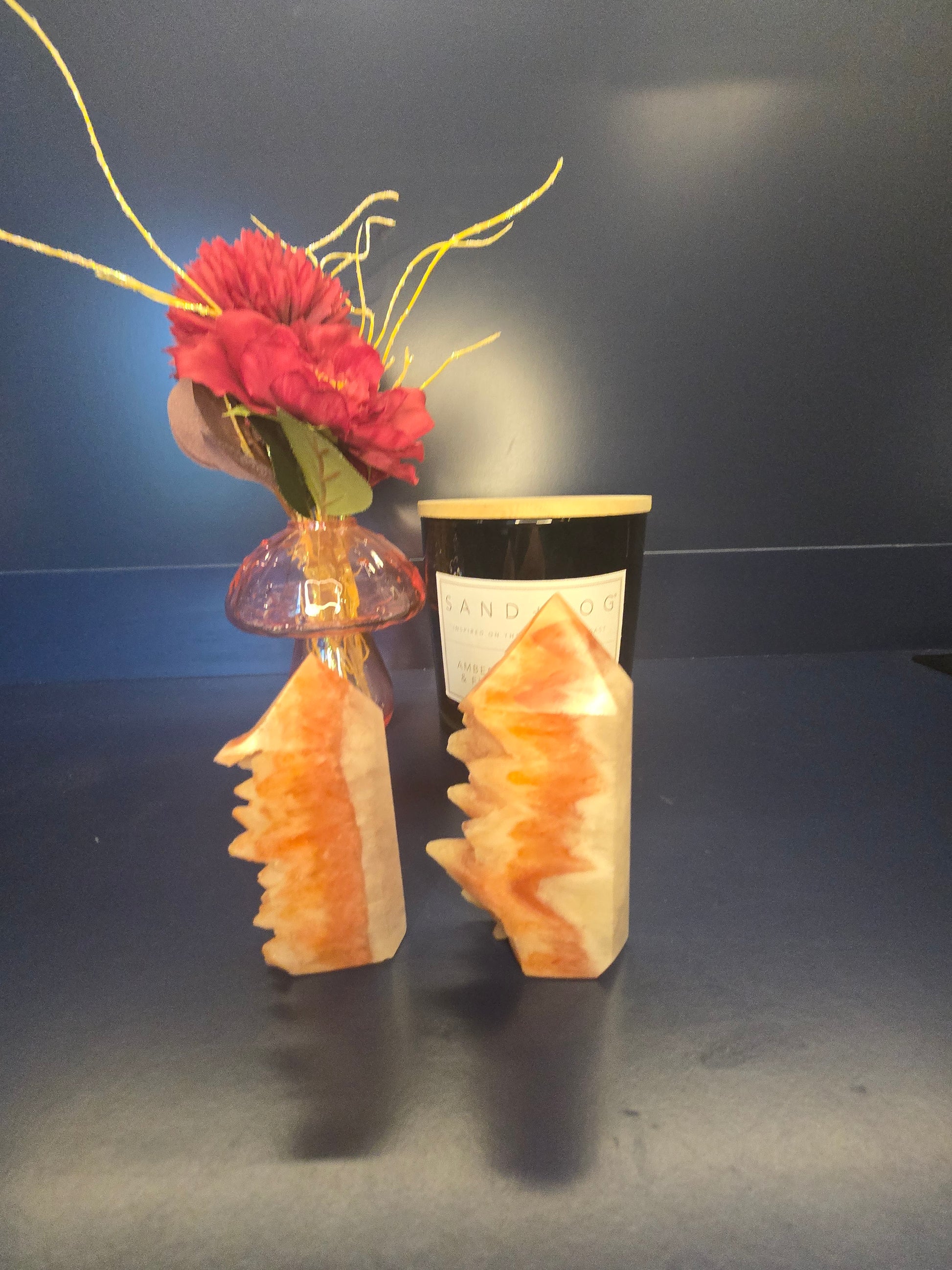 Two orange and beige dogtooth aragonite towers in front of a red flower in a vase and black candle jar