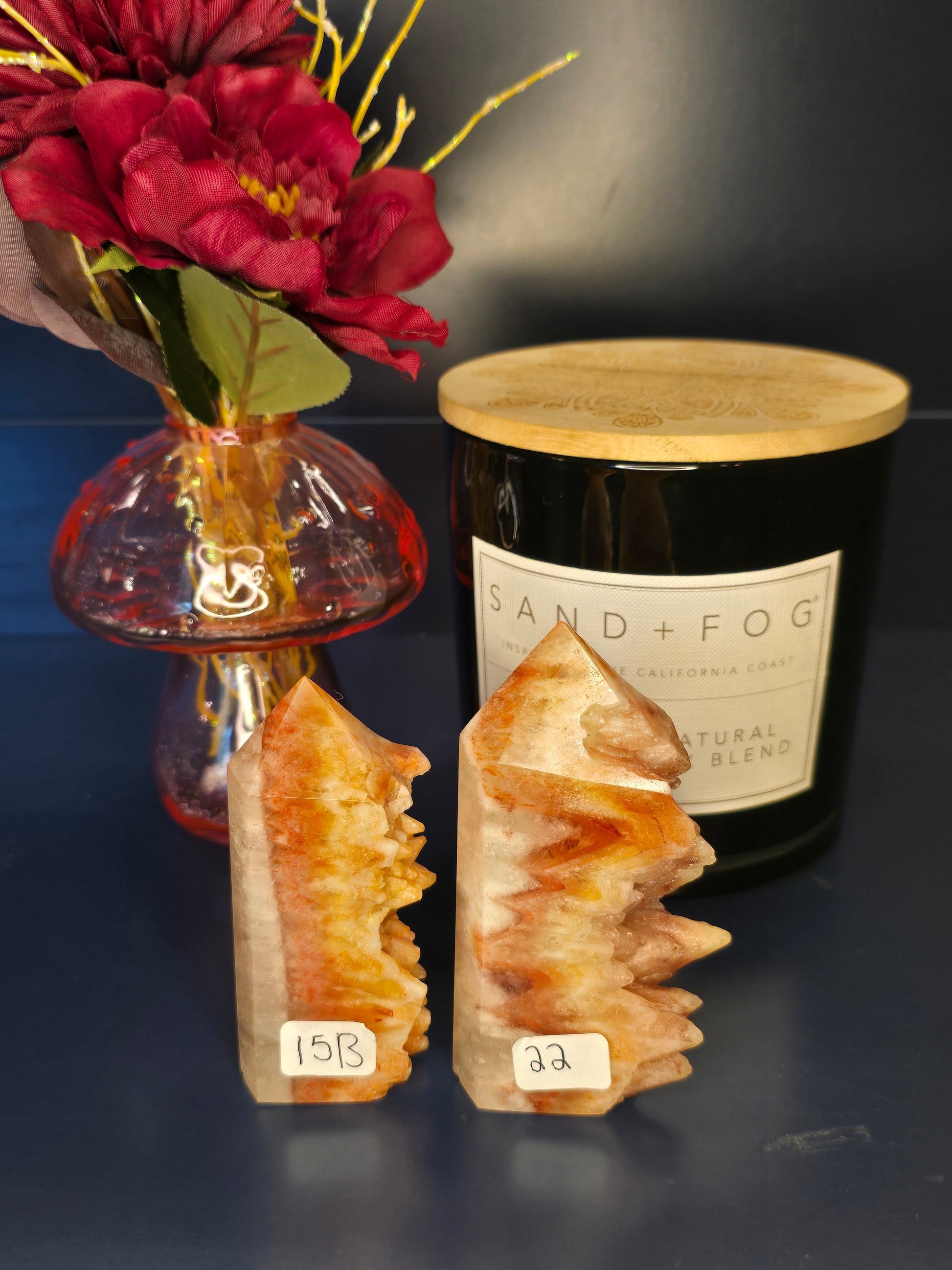 Two orange and beige dogtooth aragonite towers with numbered labels in front of a red flower in a vase and black candle jar