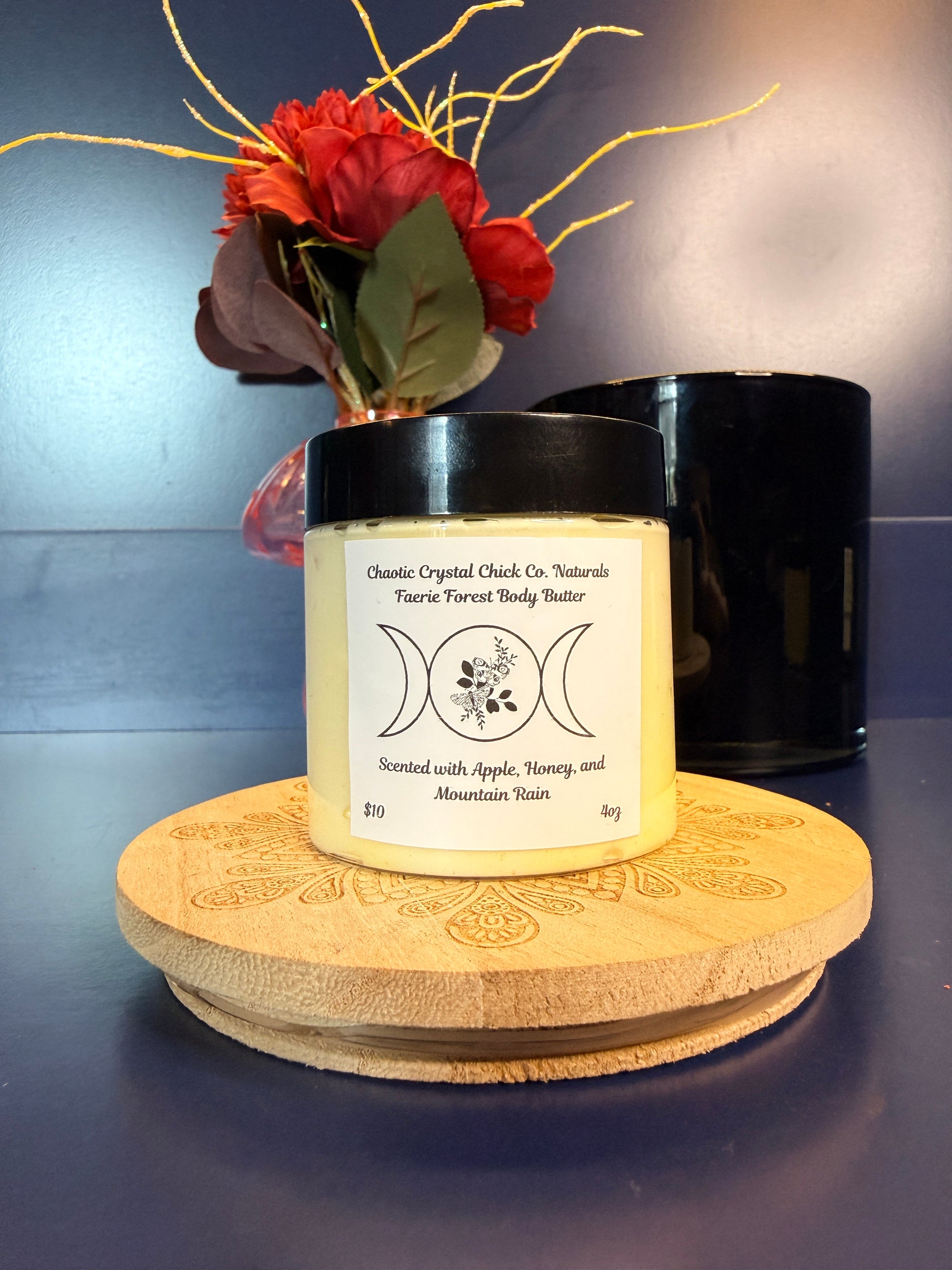 Jar of yellow body butter with a label on a decorative wooden coaster in front of a red flower and black jar