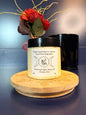 Jar of yellow body butter with a label on a decorative wooden coaster in front of a red flower and black jar