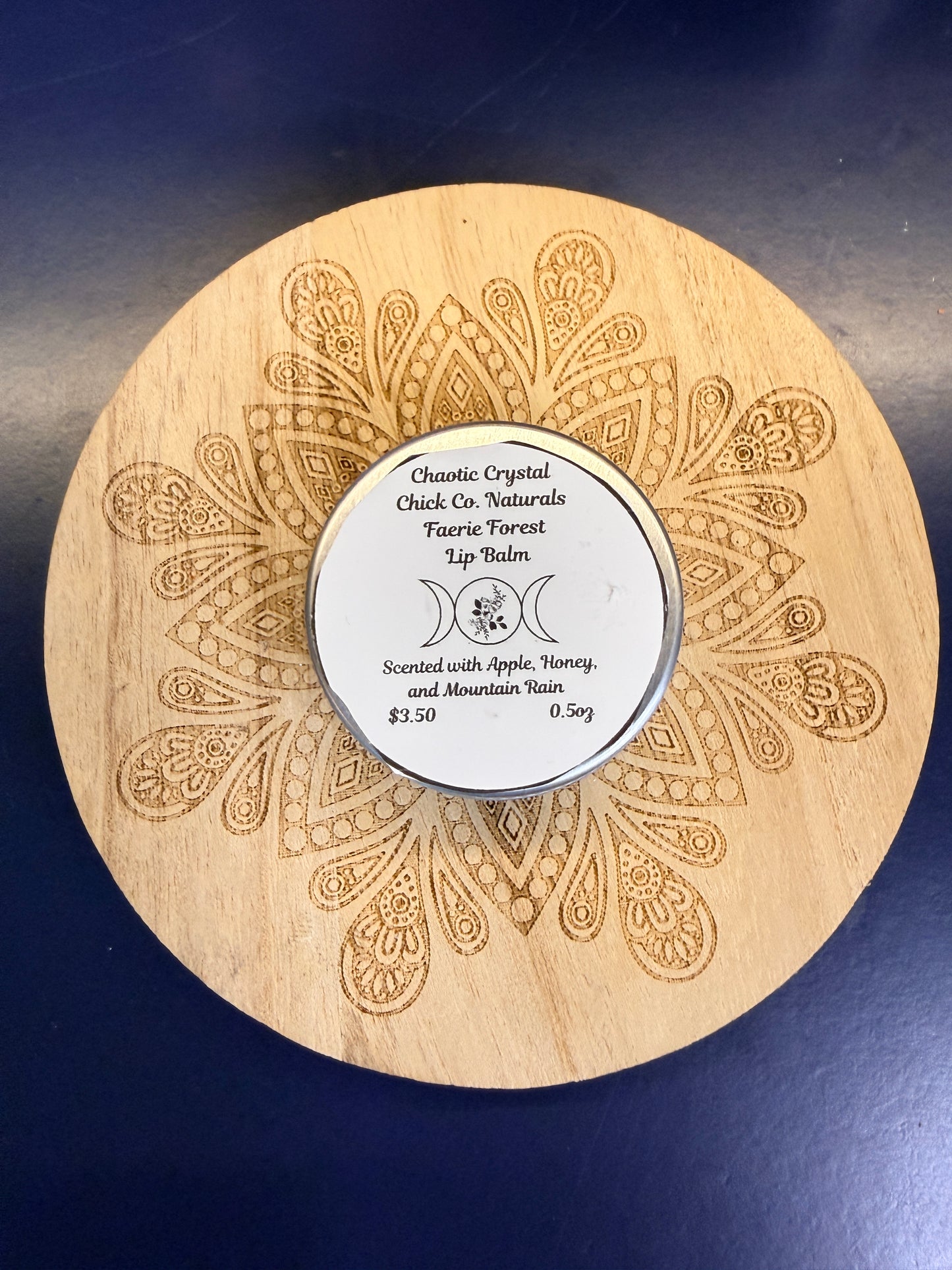Wooden coaster with intricate patterns and a round metal lip balm on a dark blue background