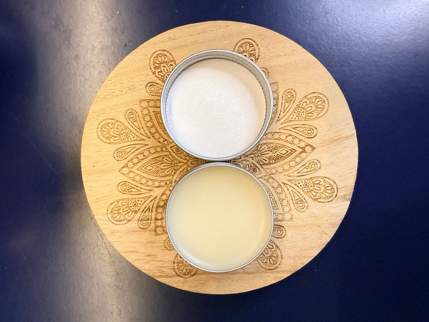 Wooden coaster with intricate patterns and a round open metal lip balm on a dark blue background