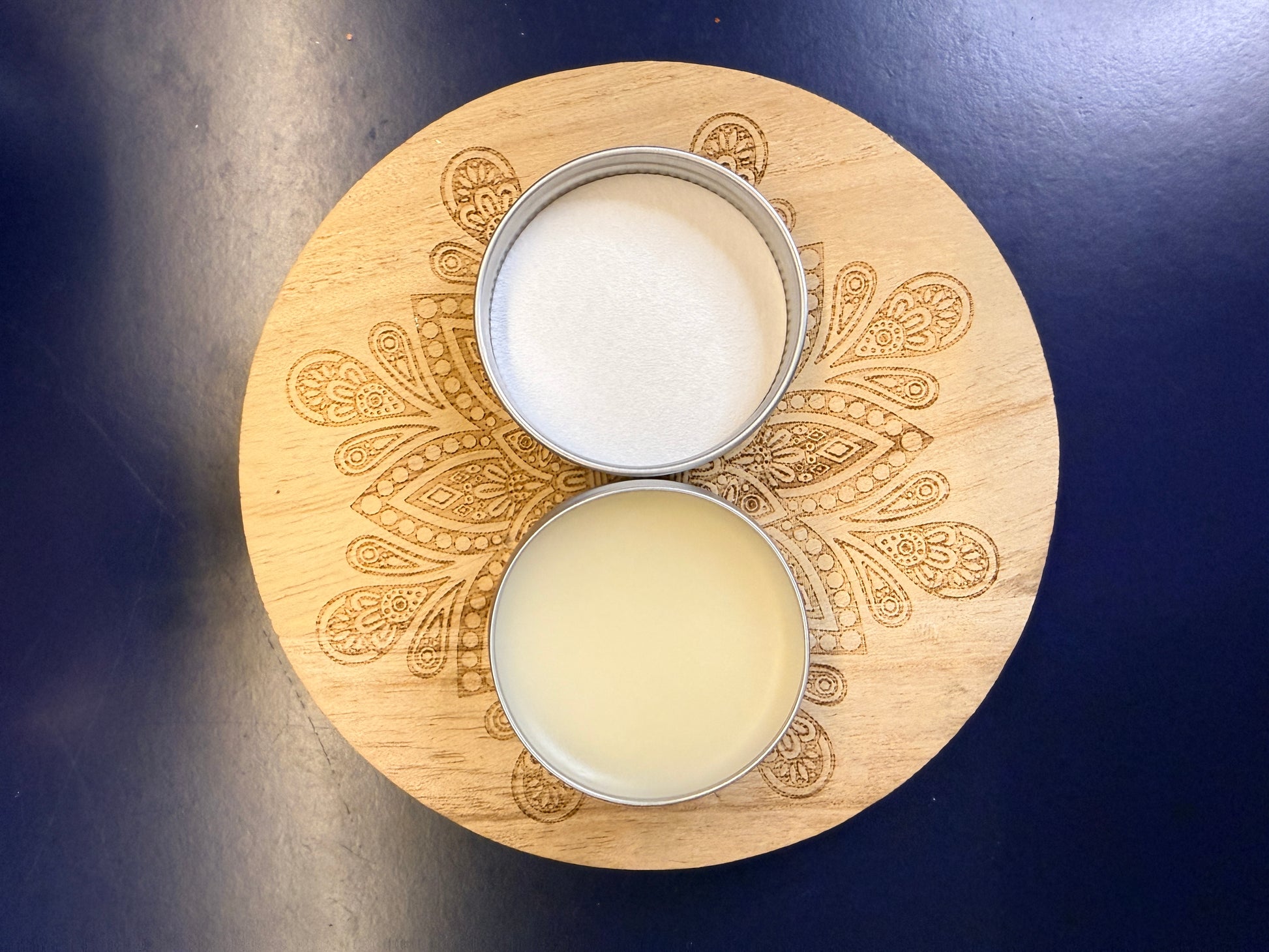 Wooden coaster with intricate patterns and a round open metal lip balm on a dark blue background