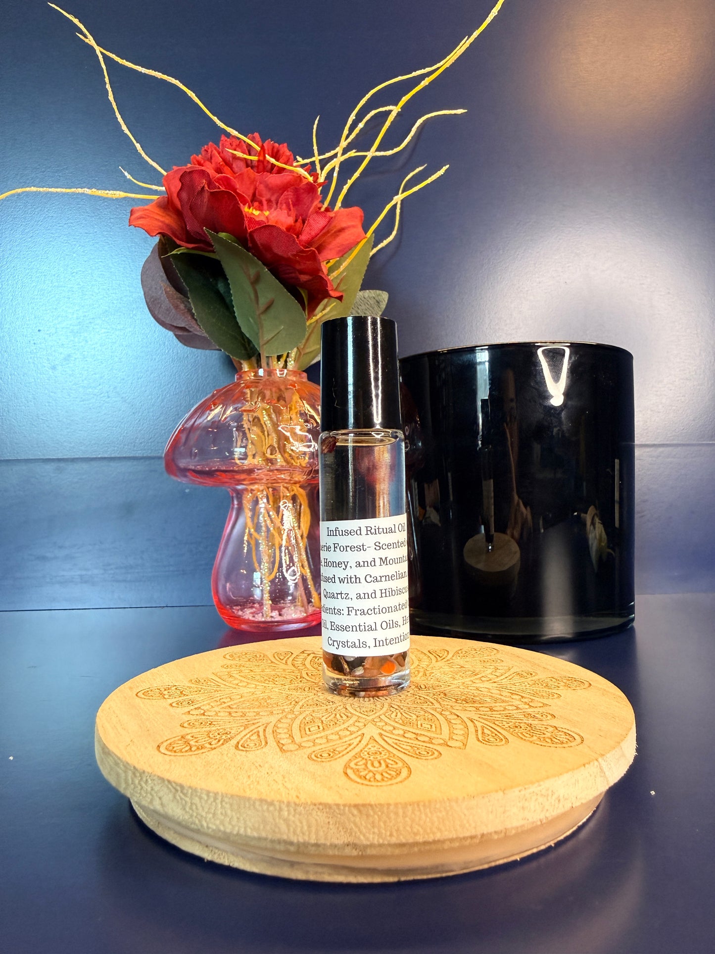 Body Oil Vial sitting on a decorative wooden coaster in front of a red flower and black jar