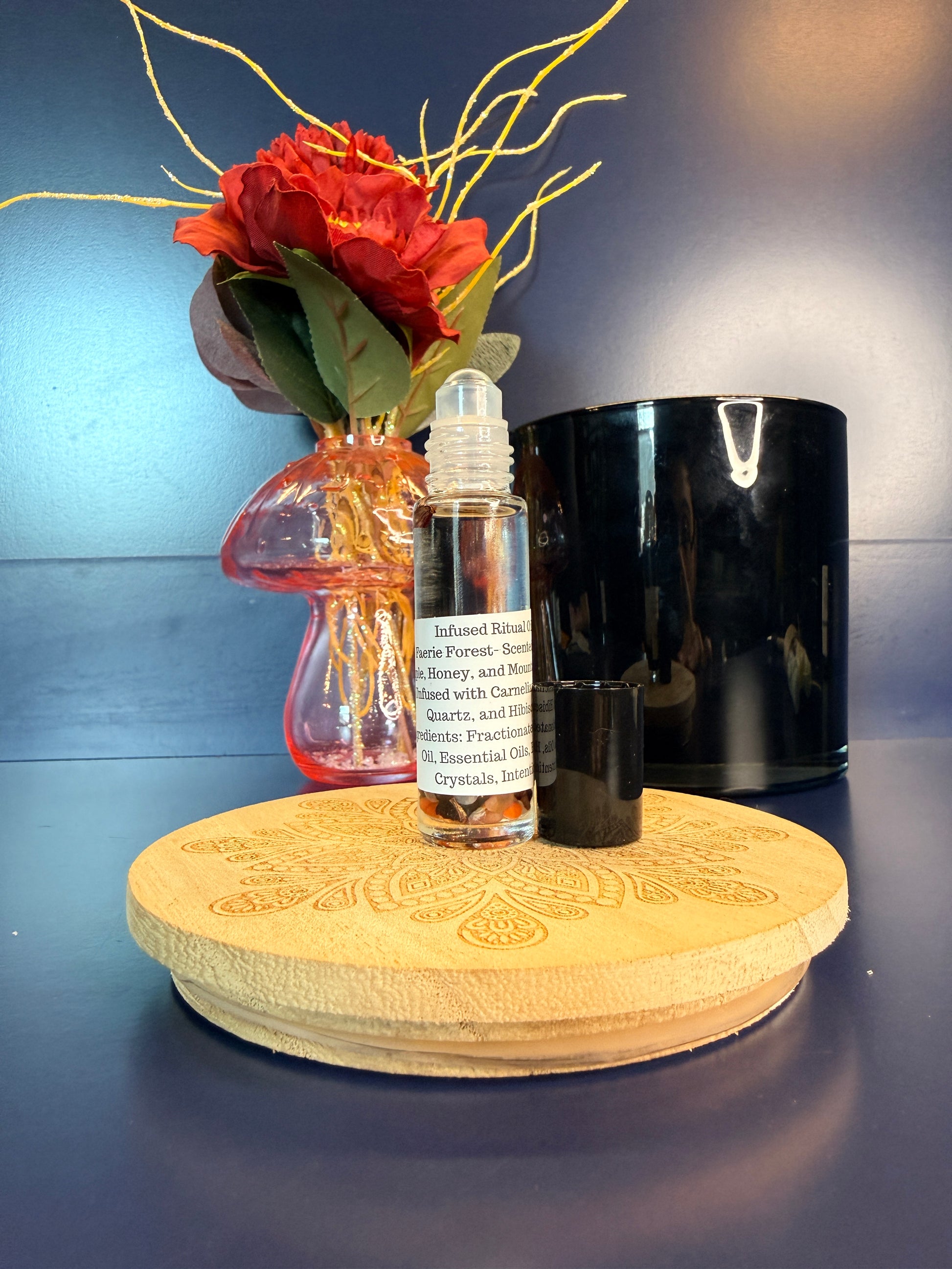 Body Oil Vial sitting on a decorative wooden coaster in front of a red flower and black jar