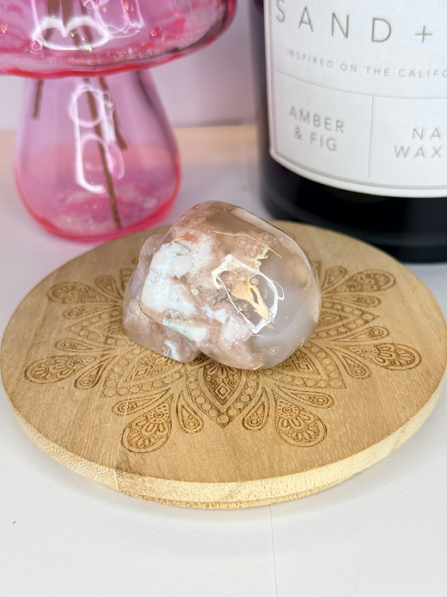 Back of a pink and white flower agate skull on a wooden coaster in front of a black candle jar and pink vase