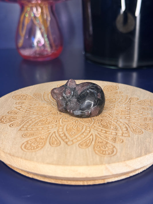 black and red garnet cat carving in front of a pink vase and black jar on a wooden coaster against a blue background