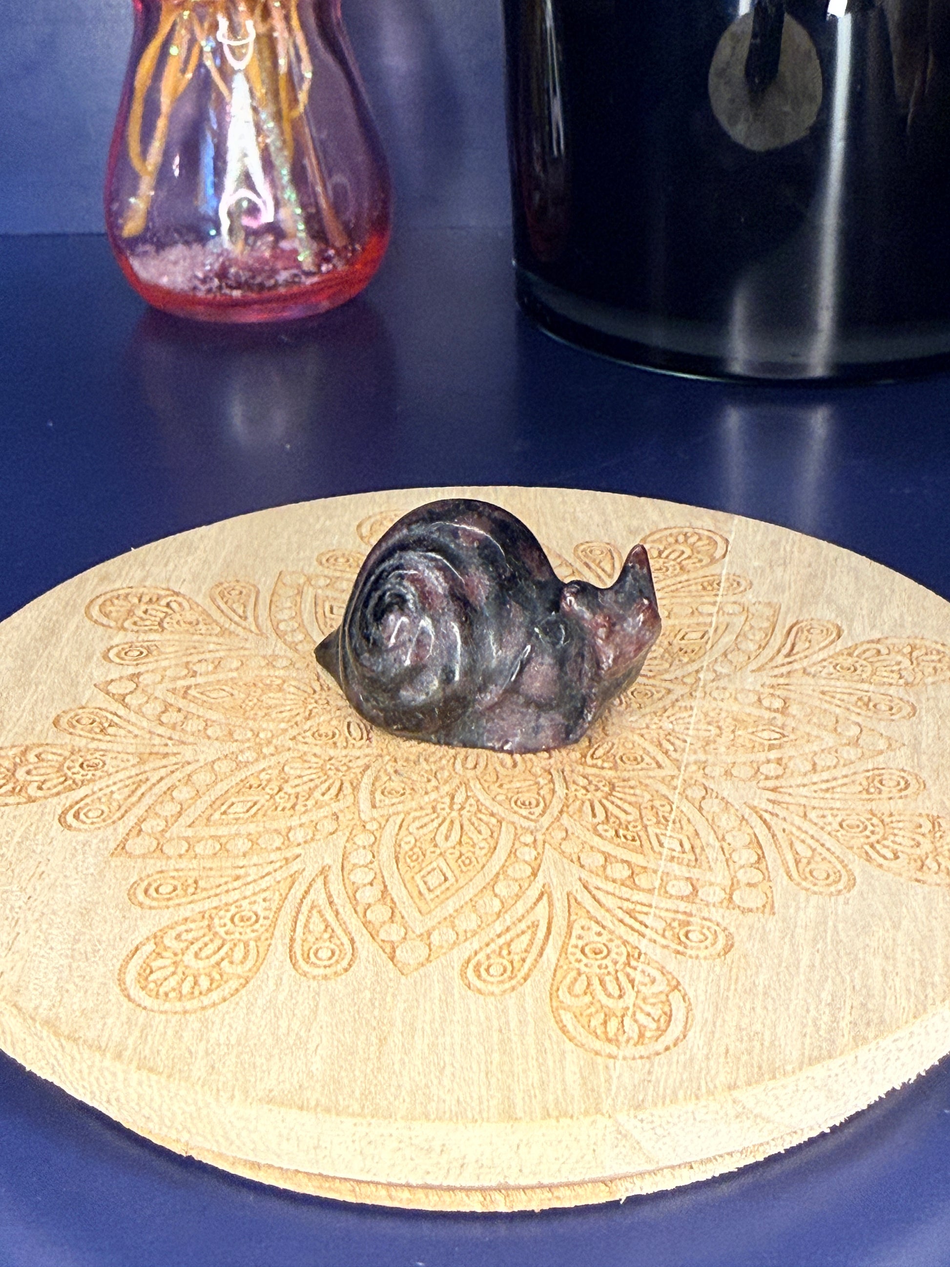 black and red garnet snail carving in front of a pink vase and black jar on a wooden coaster against a blue background