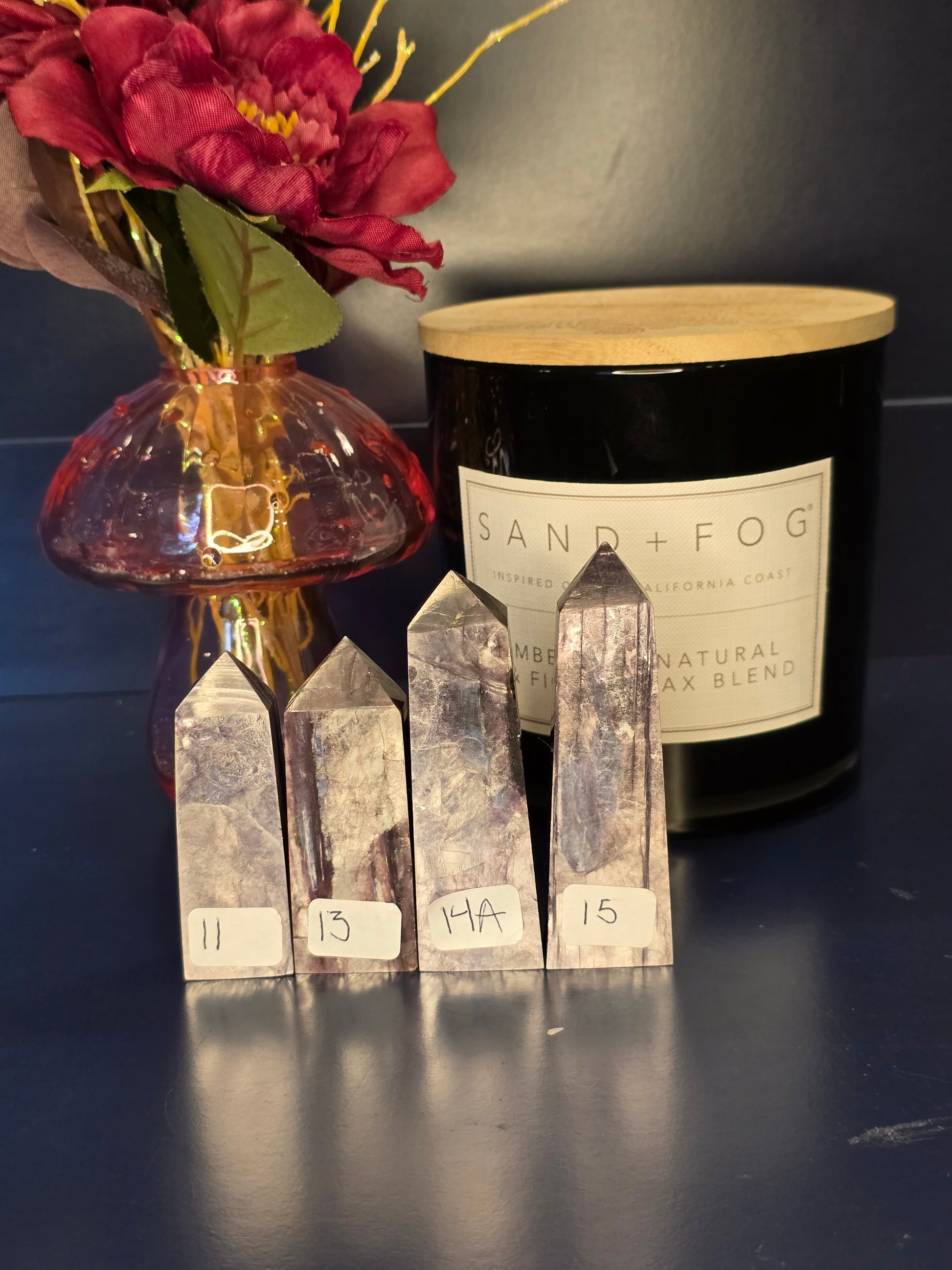 Four bright purple gem lepidolite towers with numbered labels in front of a red flower in a vase and a black candle jar