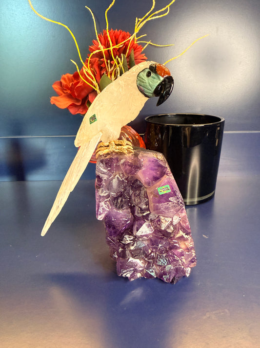 Intricately carved white gemstone parrot sitting on a purple amethyst cathedral in front of a red flower and black jar