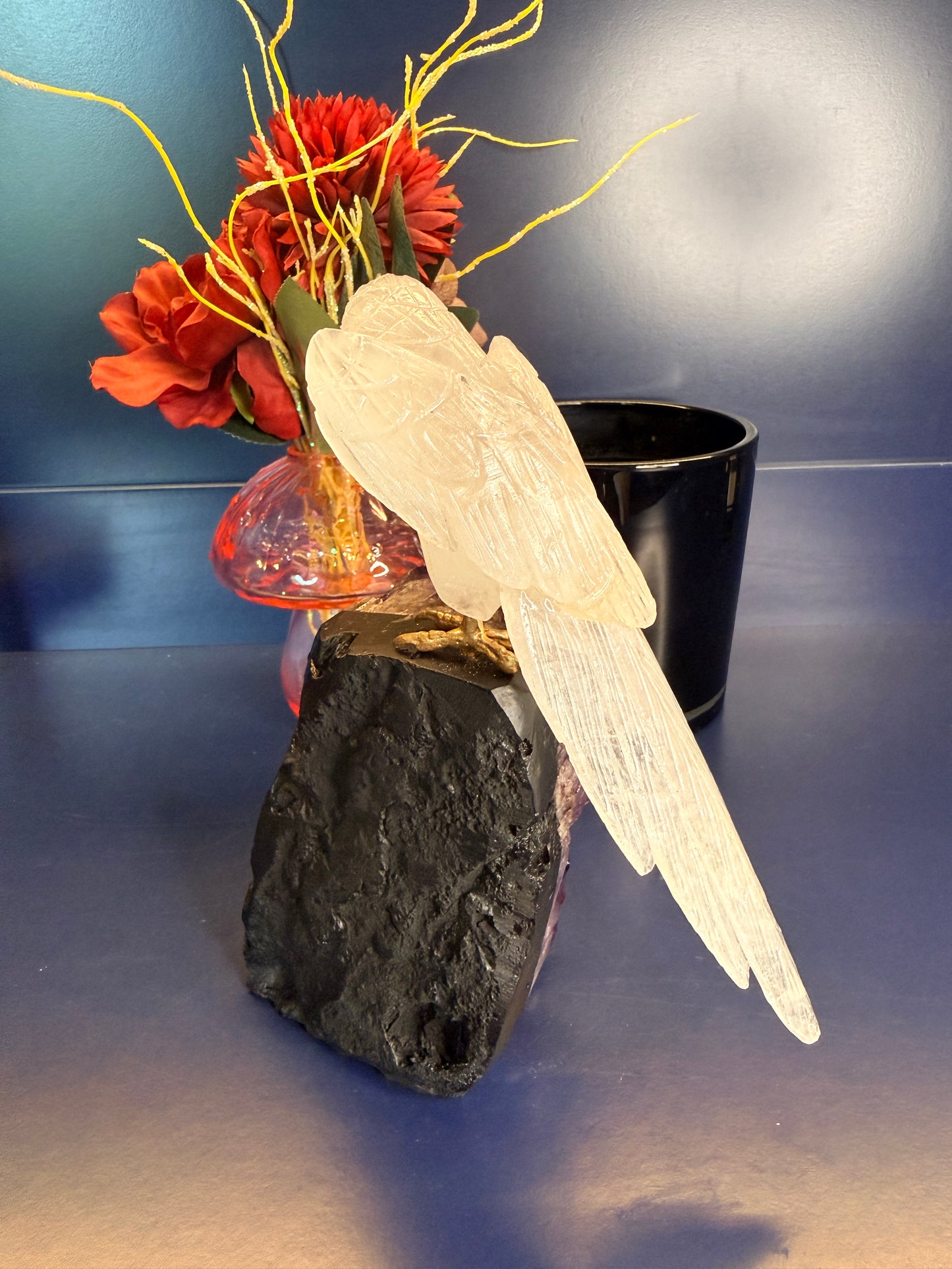 Intricately carved white gemstone parrot sitting on a purple amethyst cathedral in front of a red flower and black jar