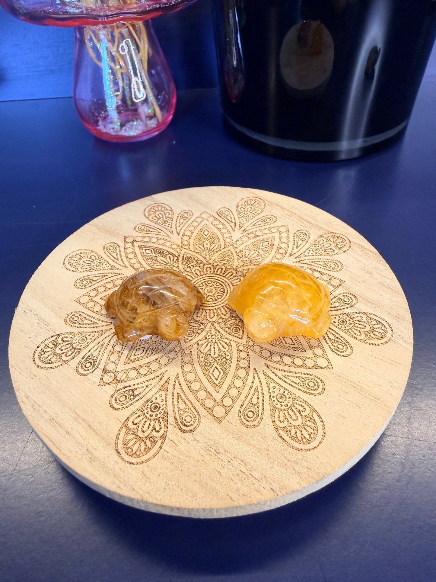 Two yellow crystal turtles on a decorative wooden coaster in front of a vase and black jar